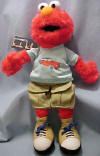 Gund Sesame Street Plush Elmo dressed in Khakis  - made from a soft plush fabric.  Elmo is wearing a light blue shirt with a car on it, khaki shorts with 2 pockets that close with velcro, and beige tennis shoes.  In 1 of the pockets is a small plush toy car.  16 inches high